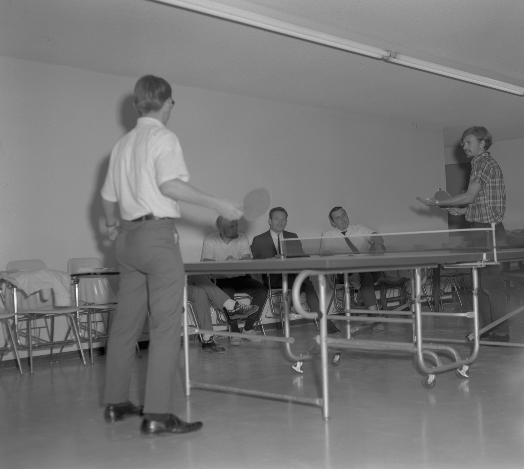 Ping pong team members battle each other as faculty look on, ca. 1967